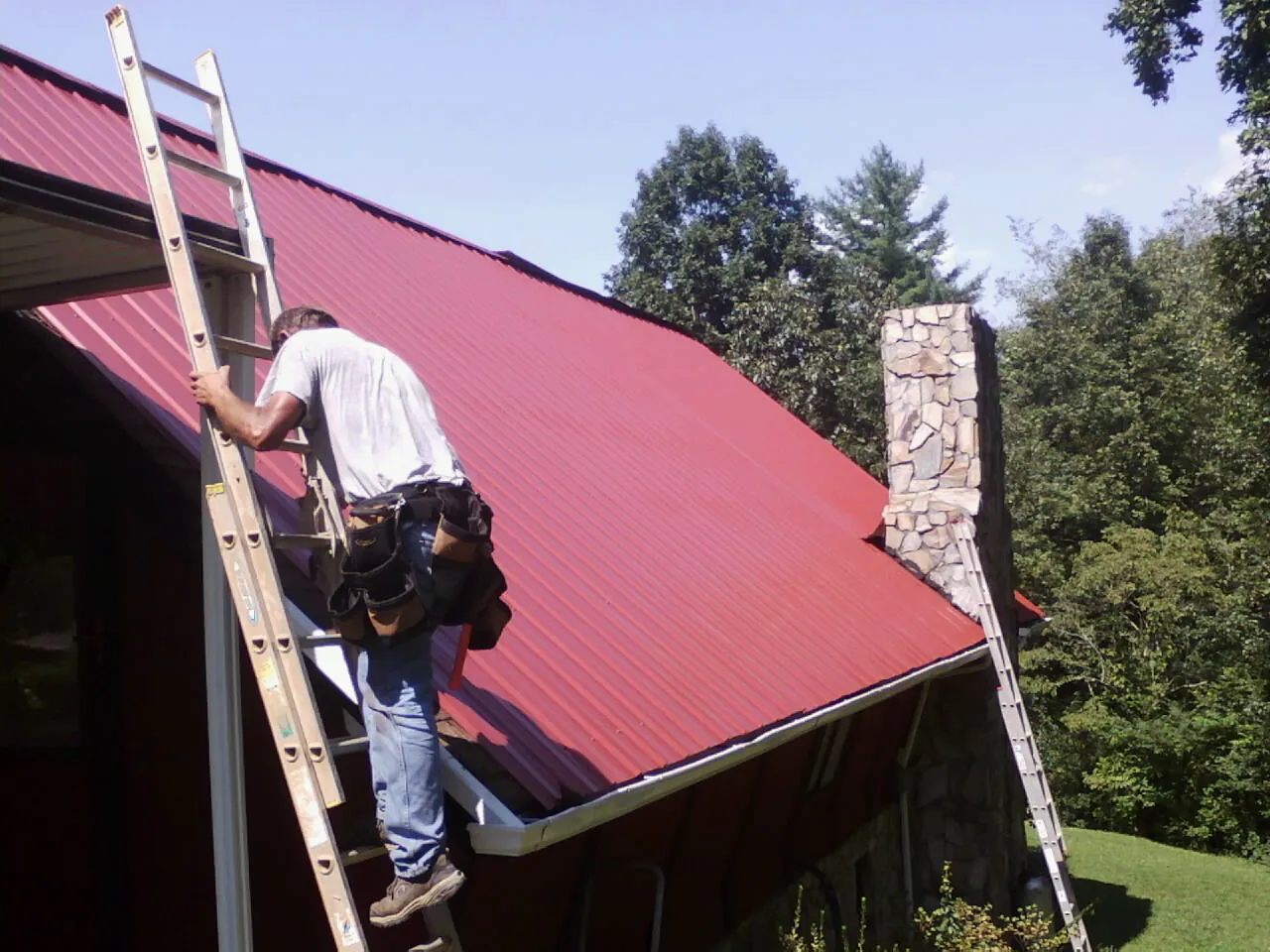 Roofer on a ladder beside a red metal roof and stone chimney — Soffit & Fascia Repair by Roofing Brothers in Elkhorn